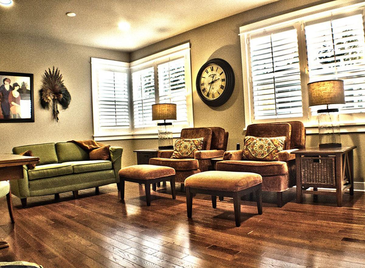 Interior view of a living area with a green sofa, two armchairs, ottomans, plantation shutters, and a wall clock.