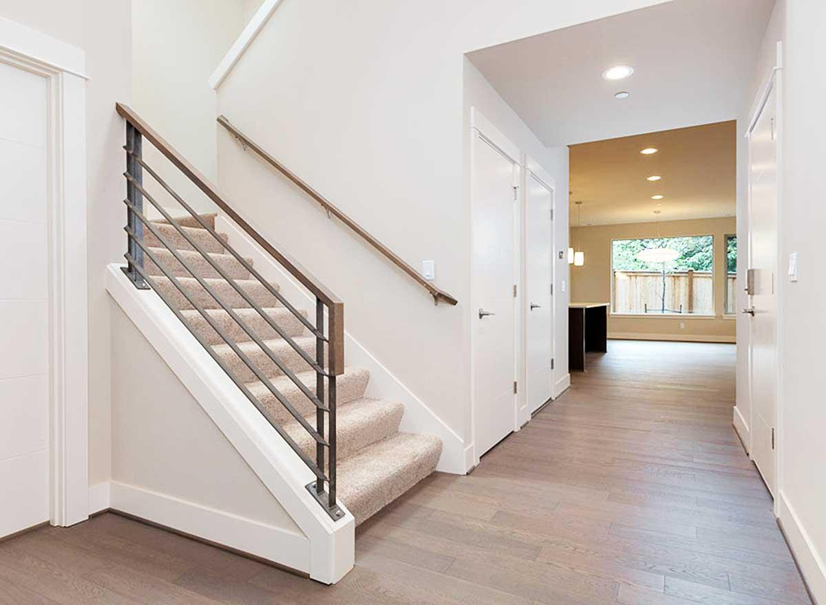 Interior view of hallway with carpeted stairs, modern metal railing, and view into a room with island and large window.