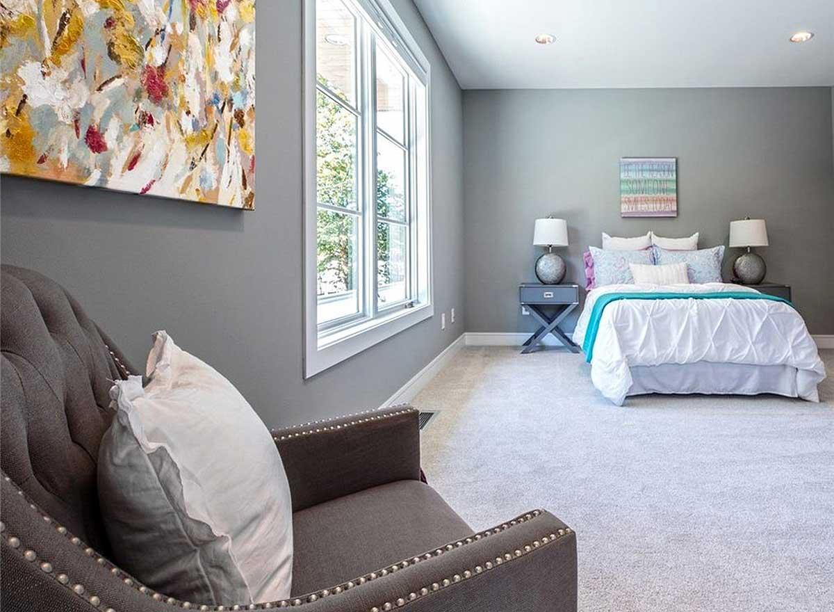 Bedroom interior with bed, nightstands, upholstered chair, and large window with white trim.