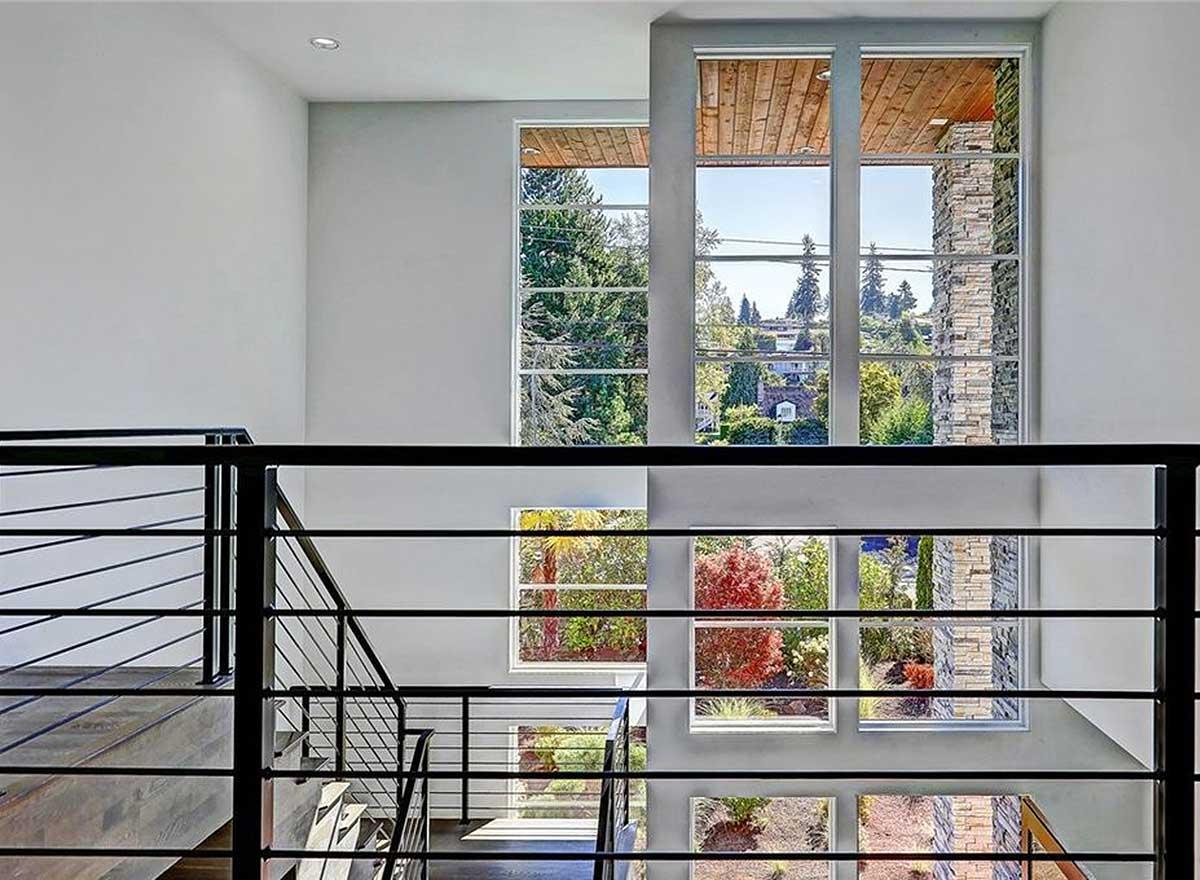 Interior view of a modern house with a multi-story staircase, black metal railings, and large windows overlooking a landscaped yard.