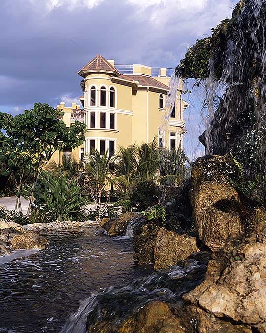Exterior of a multi-story house plan with a turret, arched windows, and a tiled roof, set against a tropical landscape with a waterfall.