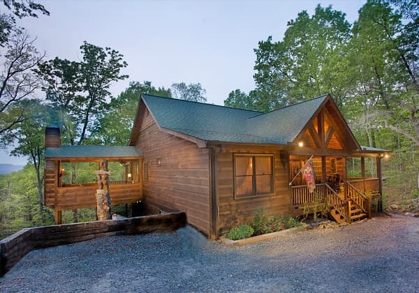 Log cabin exterior with a covered front porch, a connecting covered walkway, and a stone chimney. Features exposed timber framing under the gables.