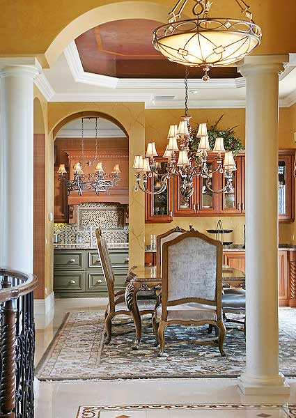Interior view of dining room with ornate chandelier, archway to kitchen with green cabinetry and tiled backsplash.
