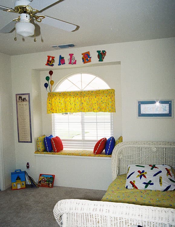 Bedroom interior with a white wicker bed, a window seat with cushions, and "HALEY" block letters on the wall.