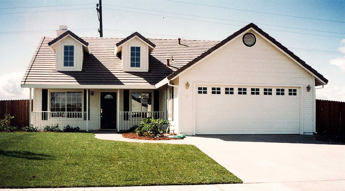 House plan exterior: one-story Ranch with front porch, dual dormers, and attached two-car garage with decorative windows.