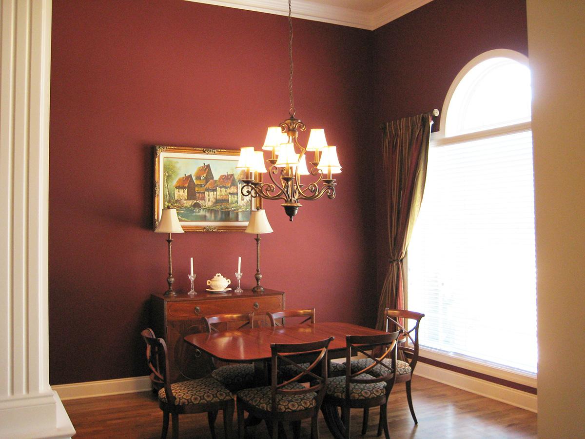 Dining room with wainscoting, traditional chandelier, and large arched window with pleated blinds and draped curtains.