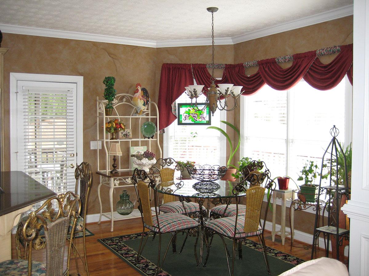 Dining area with glass tabletop, chairs, a baker's rack with decorative items, and a large window with red drapes.