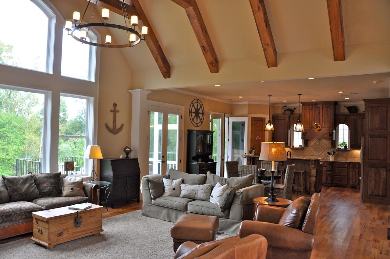 Living room with vaulted ceilings, exposed wood beams, large windows, fireplace, and open-concept kitchen.