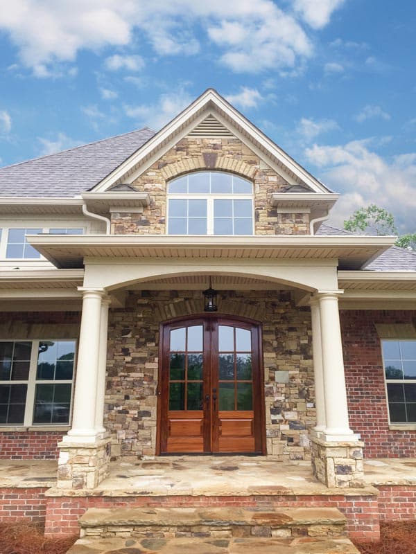 House plan exterior with arched stone entryway, double wood doors, brick facade, and classical columns supporting a covered porch.