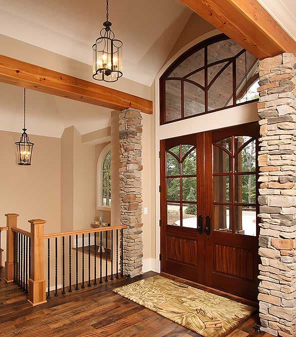 Interior view of a grand entrance with double wooden doors, stone pillars, exposed beams, and pendant lighting.