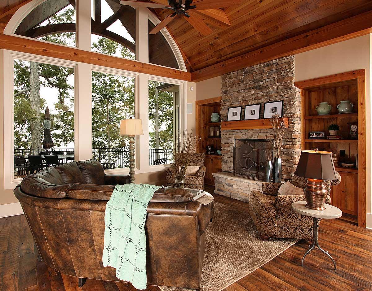 Great room with vaulted wood ceiling, stone fireplace, built-in bookshelves, and large windows overlooking trees and water.