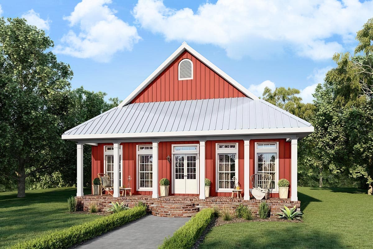 A red house with a white metal roof, porch, and columns. The house has a brick foundation, a walkway, and sits among trees under a blue sky.