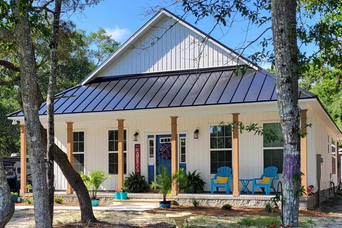 White farmhouse with a black roof and blue door. Porch has blue chairs and potted plants. Trees frame the house on a sunny day.