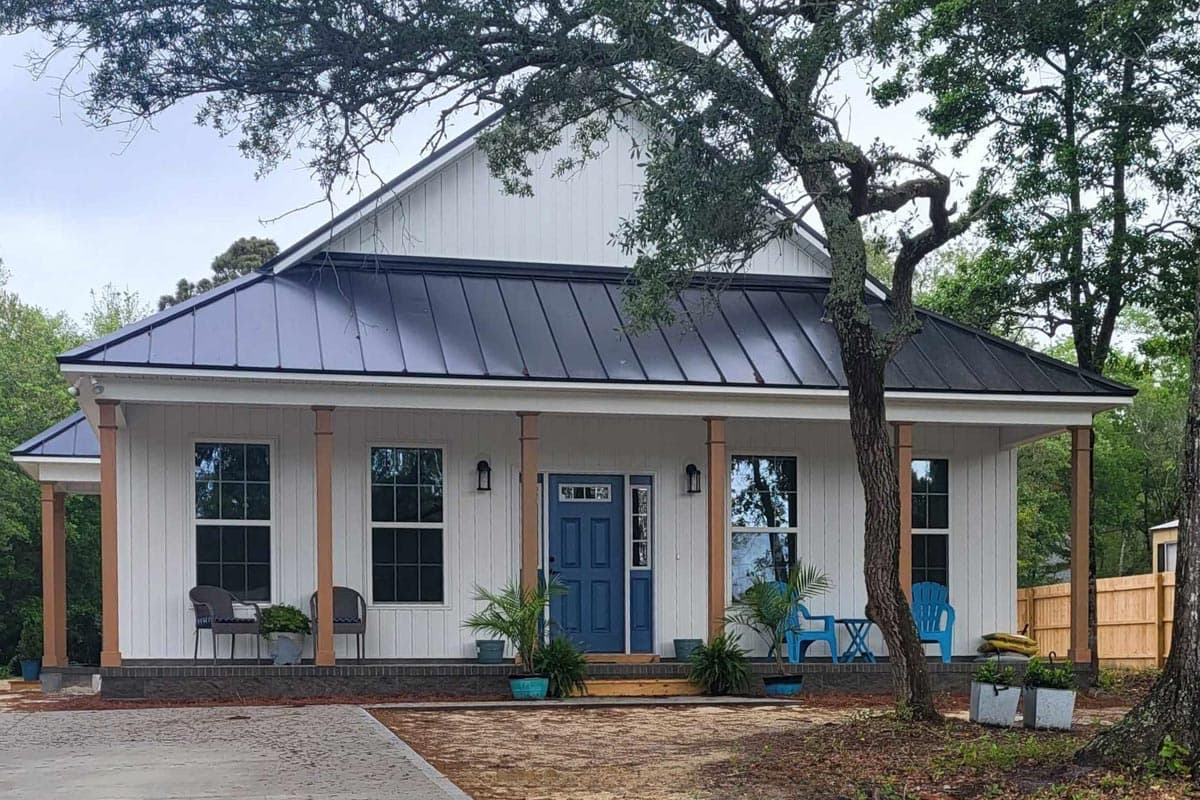 A white house with a dark metal roof, blue door, and porch. Plants and blue chairs decorate the porch, with trees framing the house.