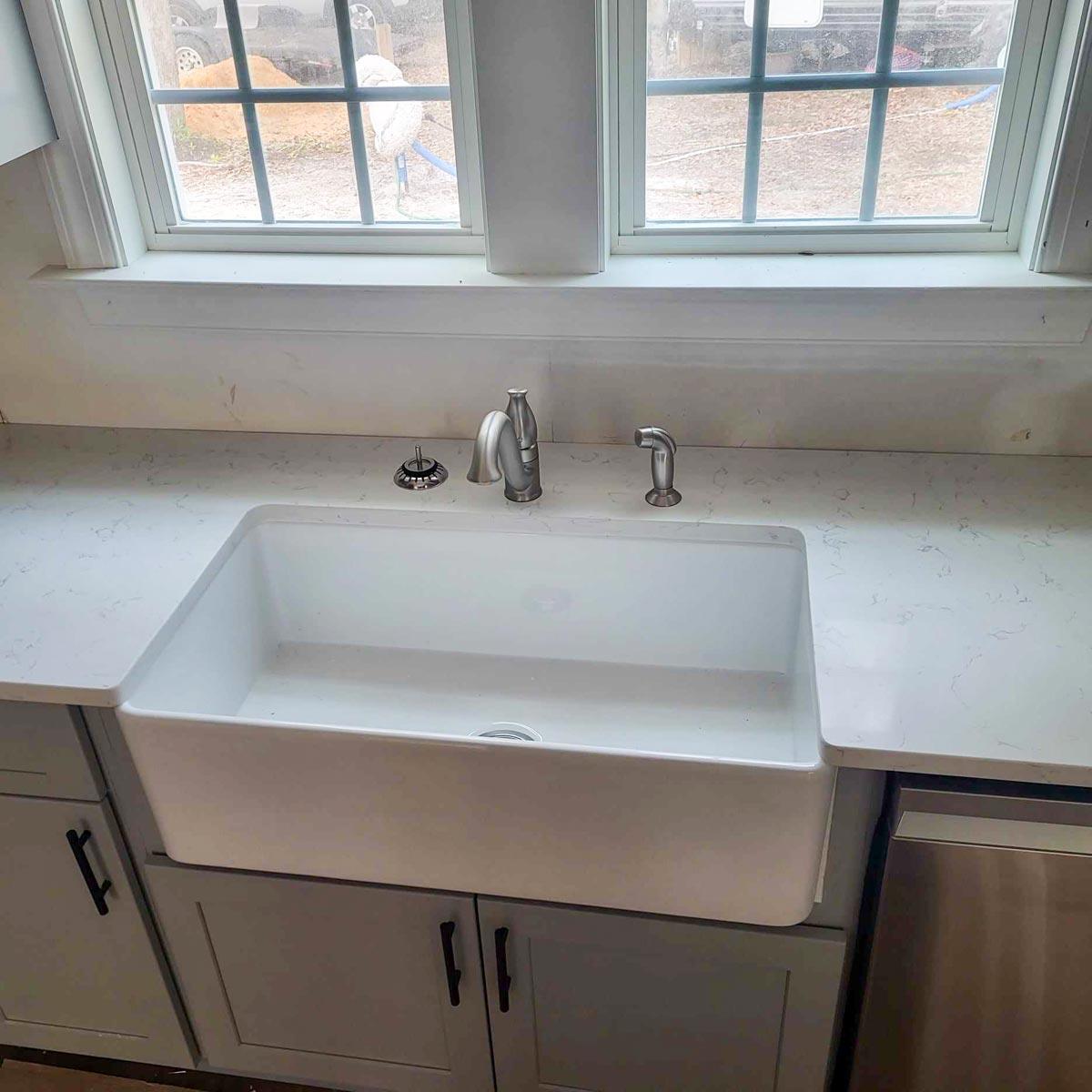 A farmhouse kitchen sink is set into white countertop. Gray cabinets are below, with a chrome faucet and two windows overhead providing natural light.