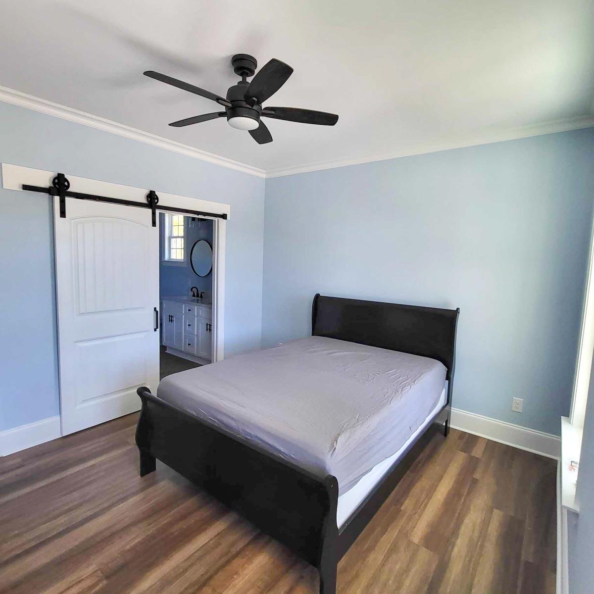 Bedroom with blue walls, dark bed frame, and ceiling fan. A sliding barn door leads to a glimpse of a bathroom. Wooden floor throughout.