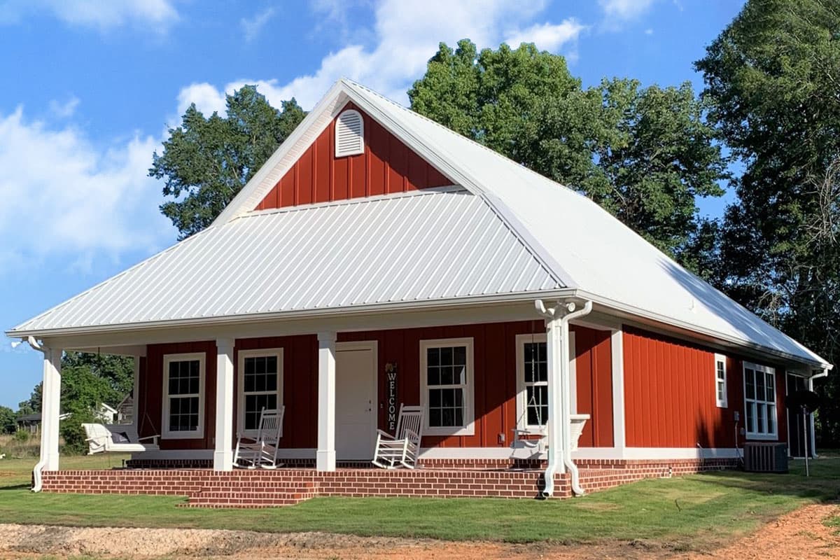 A red house with a white metal roof and porch, two rocking chairs, and a welcome sign. A swing hangs from the porch. Blue sky with trees.