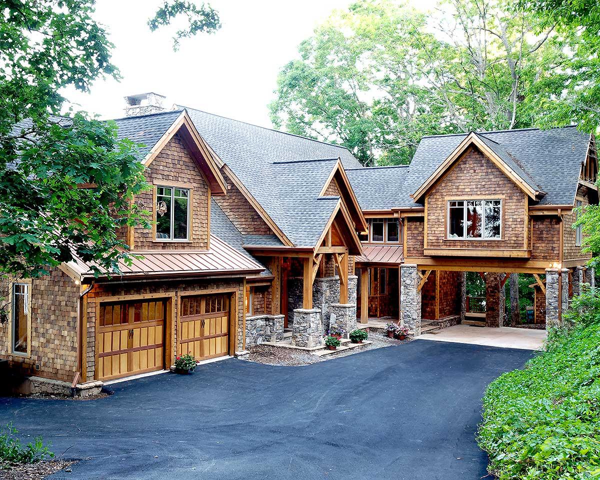 House plan exterior: Two-story mountain lodge with wood shake siding, stone-clad columns, multiple gables, and a drive-through porte-cochère.