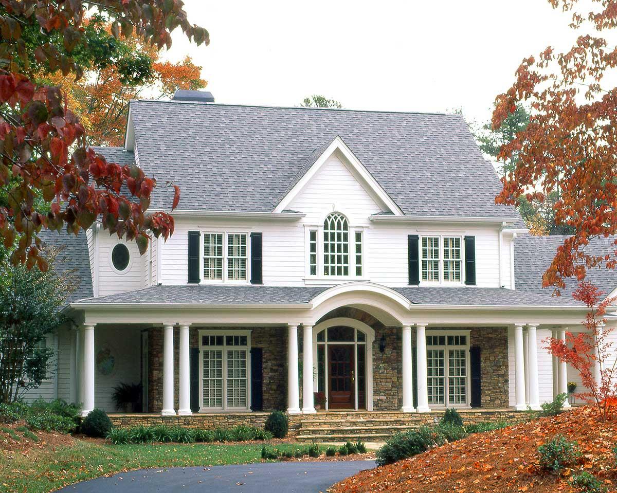 House plan exterior: two-story white home with stone base, arched entry, wraparound porch, columns, and gables.