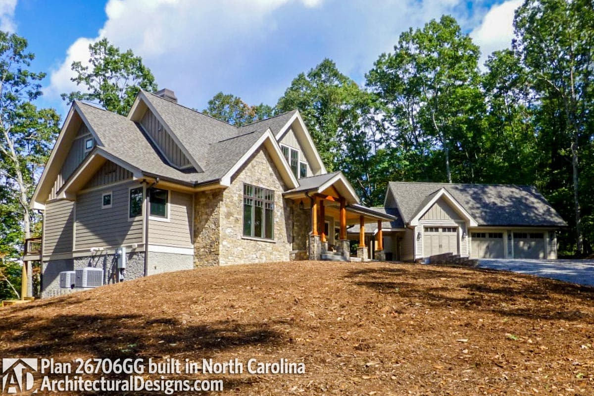House plan exterior featuring stone facade, gabled dormers, covered porch with timber posts, and three-car garage.