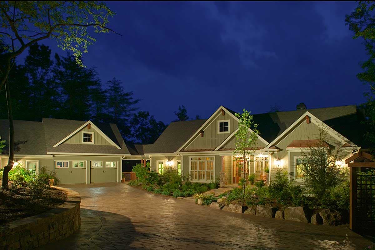 House plan exterior. Craftsman style with gabled roof, dormers, and attached garage. Stone retaining wall and paved driveway lead to the entrance.