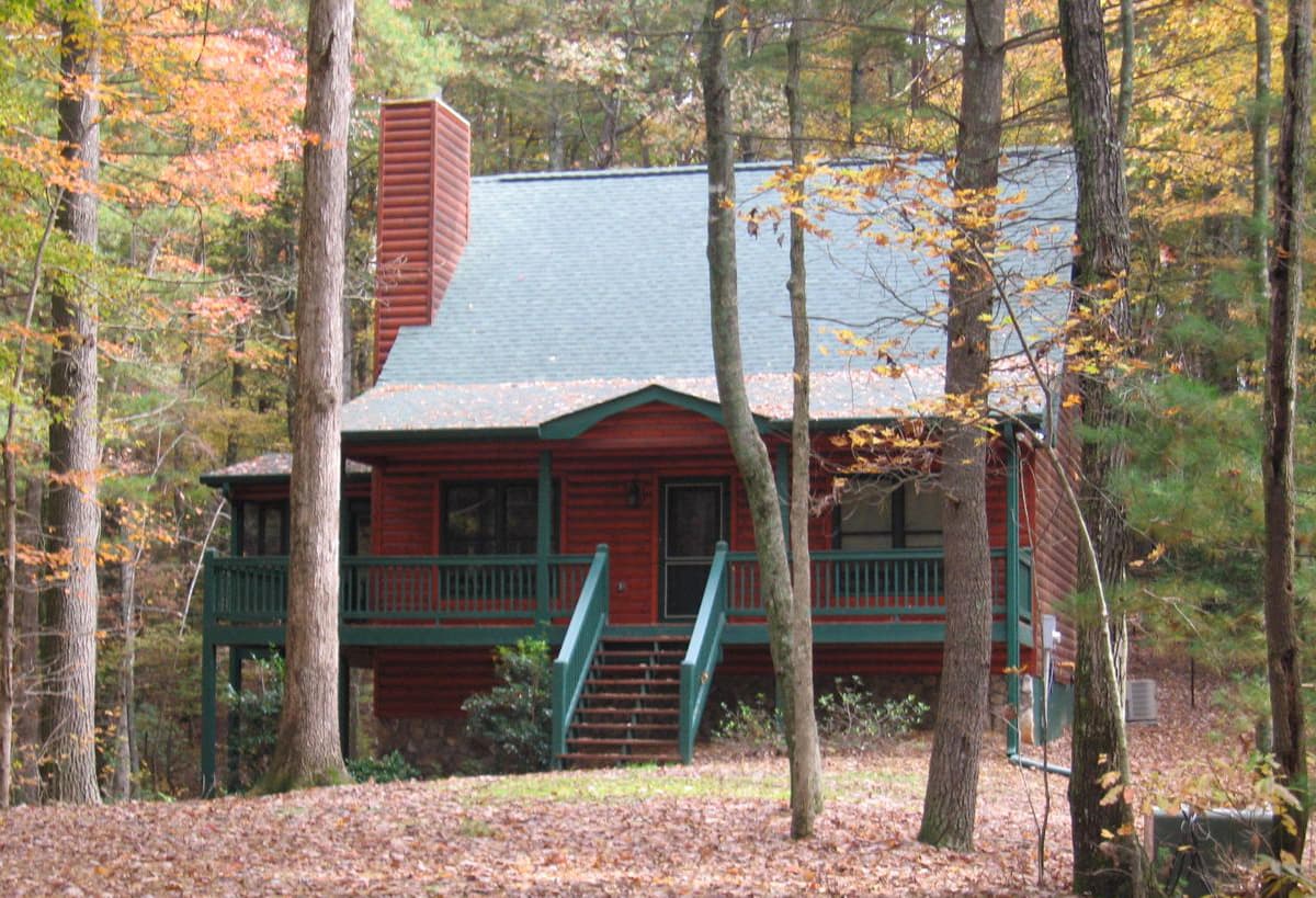 Log cabin exterior with a red exterior, green trim, wraparound porch, and prominent stone chimney.
