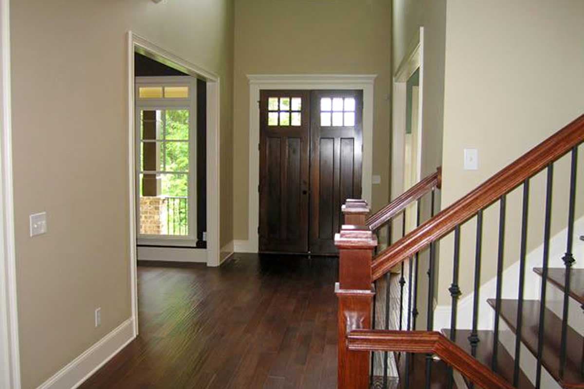 Interior view of a house foyer with double wooden doors, a staircase with wood railings, and a doorway to another room with a large window.