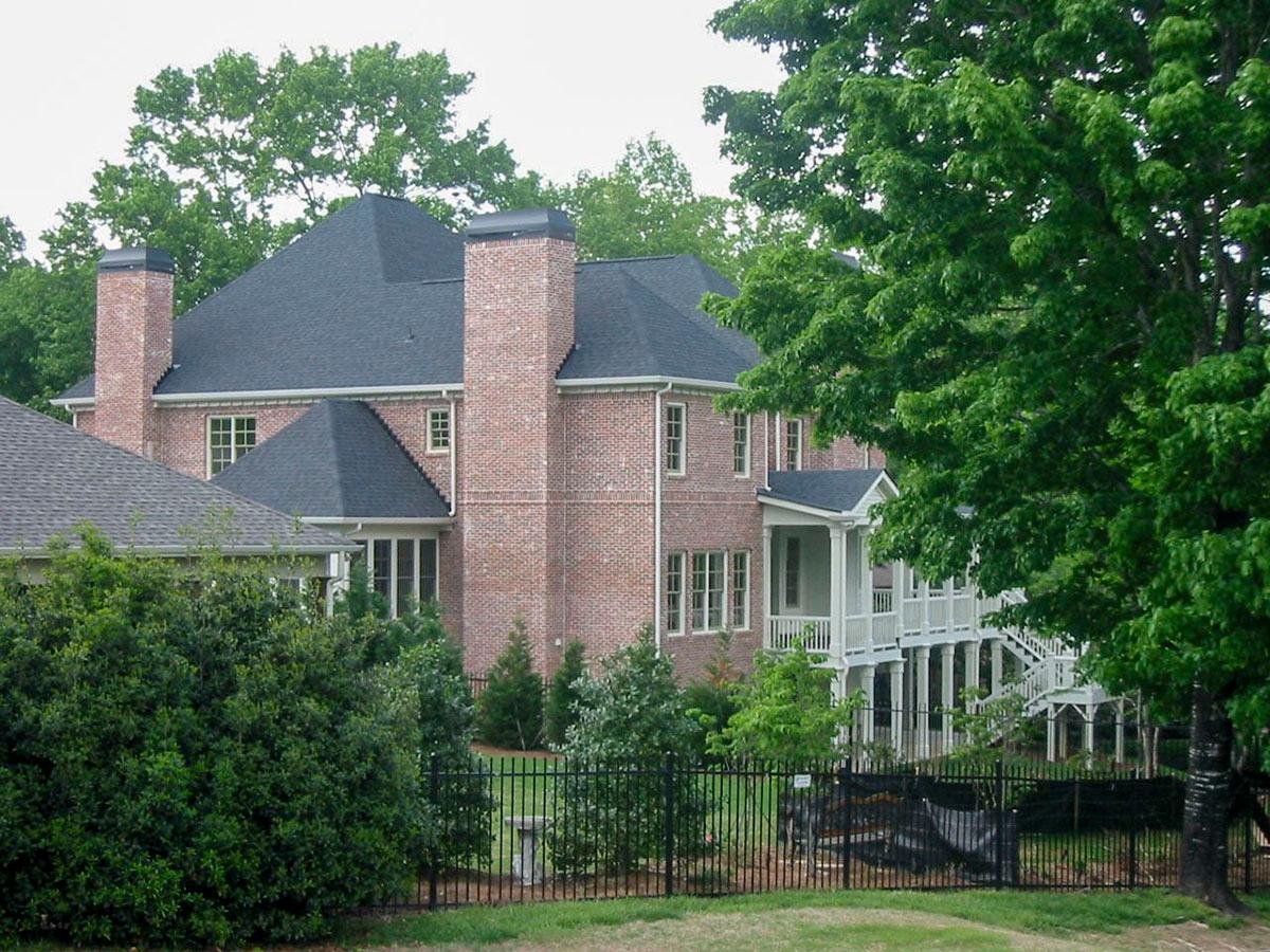 House plan exterior: brick two-story with dark roof, multiple gables, prominent chimneys, and a wraparound porch with columns.