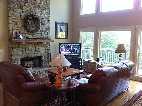 Living room interior with stone fireplace, leather sofas, television, and large windows overlooking wooded area.