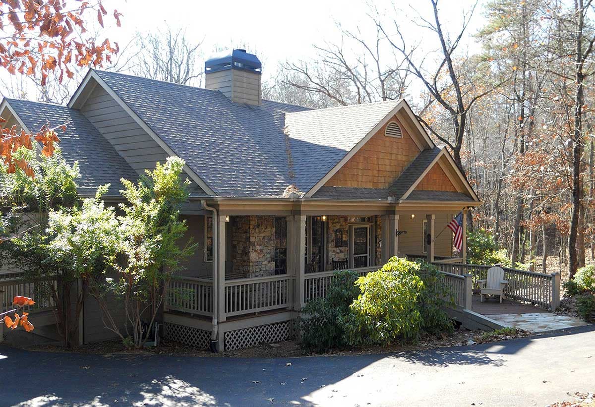 House plan exterior featuring a wraparound porch with stone accents, gables with wood shingles, and a prominent chimney.