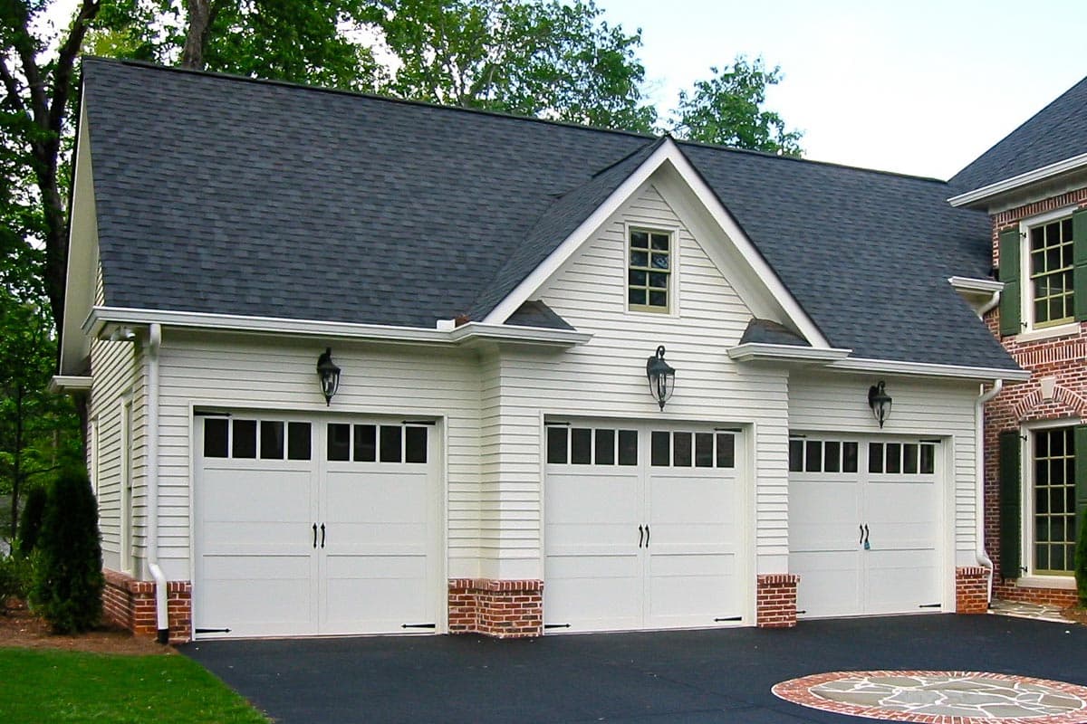 Three-car garage exterior with white siding, brick accents, gable dormer with window, and dark roof.