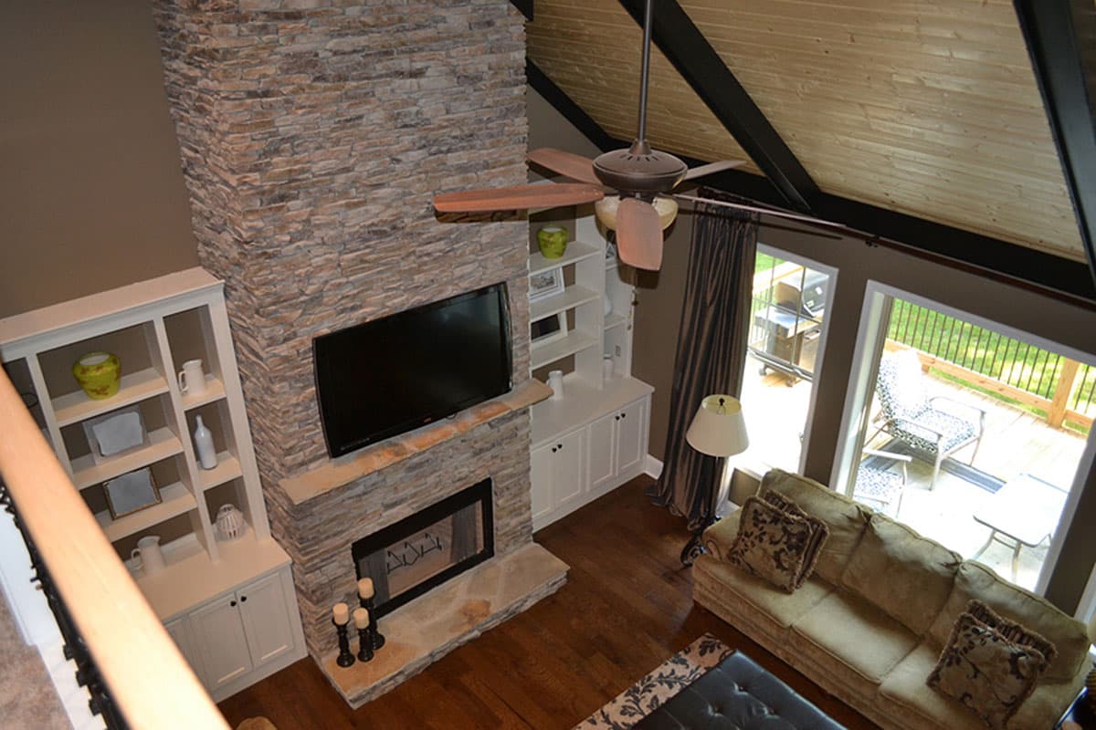 Great room interior with stone fireplace, mounted TV, built-in shelving, vaulted wood ceiling, and deck access.