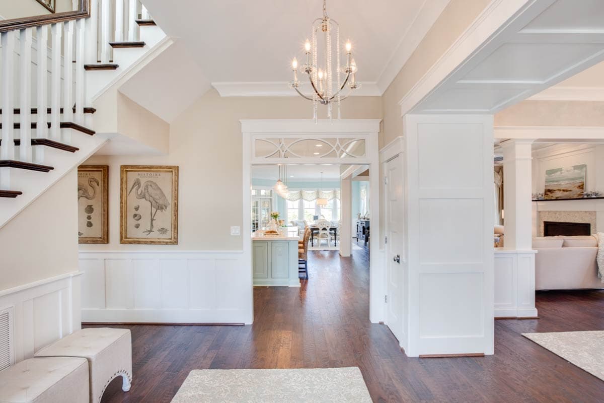 Interior view of a house entryway with a staircase, wainscoting, a chandelier, and views into the kitchen and living room.