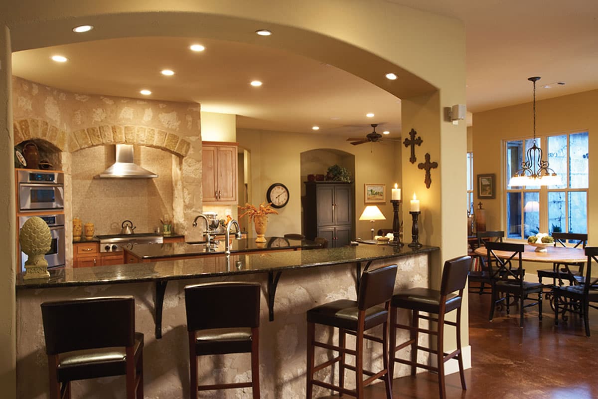 Kitchen interior with island seating, granite countertops, stainless steel range hood, and arched stone surround. Dining area visible.