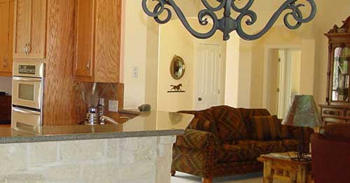 Interior view of kitchen area with wood cabinets, double oven, granite countertop, and living room with sofa and decorative chandelier.