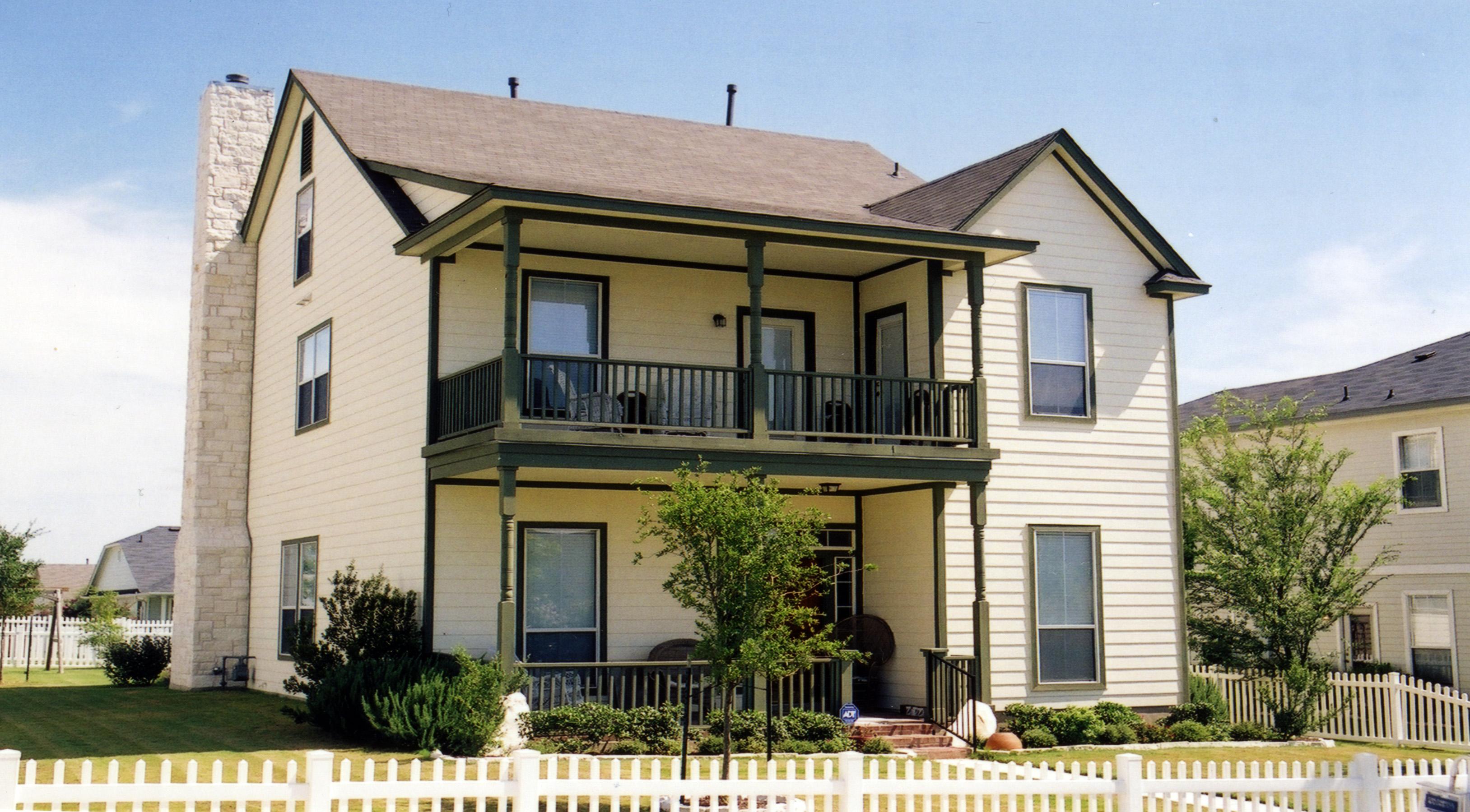 Two-story house plan exterior with a prominent stone chimney, wraparound porches on both levels, and a gable roof.