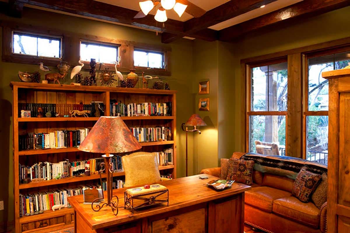 Rustic home office with wooden beams, built-in bookshelves, desk, leather sofa, and decorative lamps.