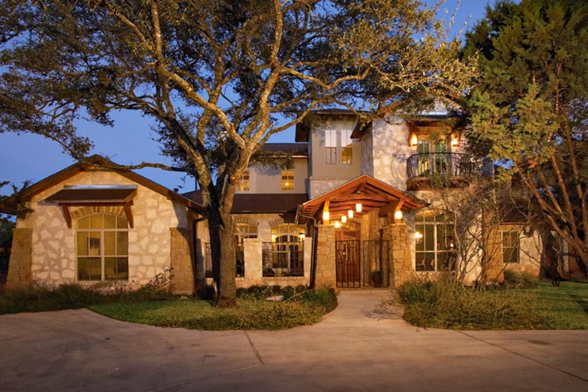 House plan exterior featuring stone facade, covered entrance with arched windows, gables, and a second-story balcony.