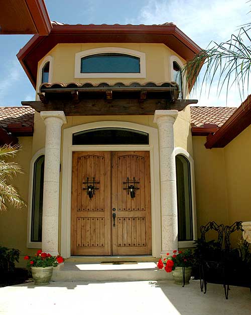 House plan exterior featuring double wooden doors with wrought iron, stone columns, arched windows, and a tiled roof.