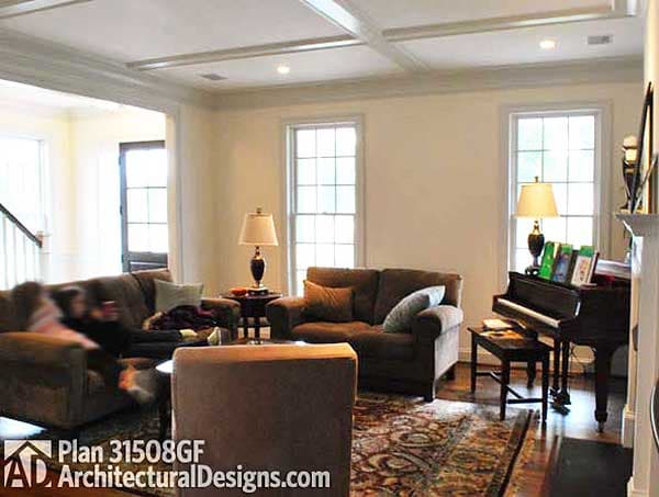 Interior view of living room with coffered ceiling, two windows, seating area, piano, and fireplace.