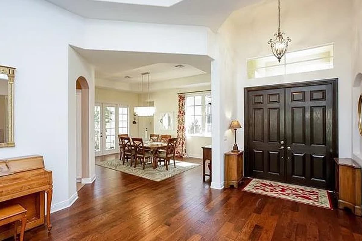 Interior view of house entryway with double dark wood doors, leading to a dining room with wood table and chairs, and a grand chandelier.