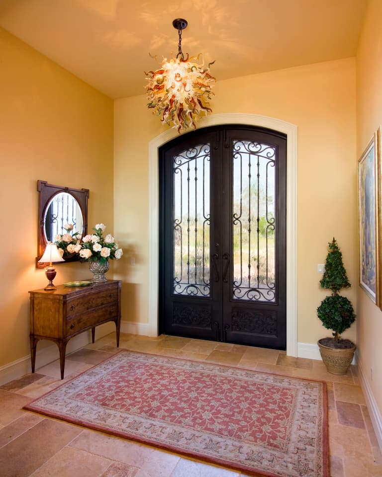Entryway with double wrought iron doors, ornate chandelier, antique console table with mirror and floral arrangement, and patterned rug.