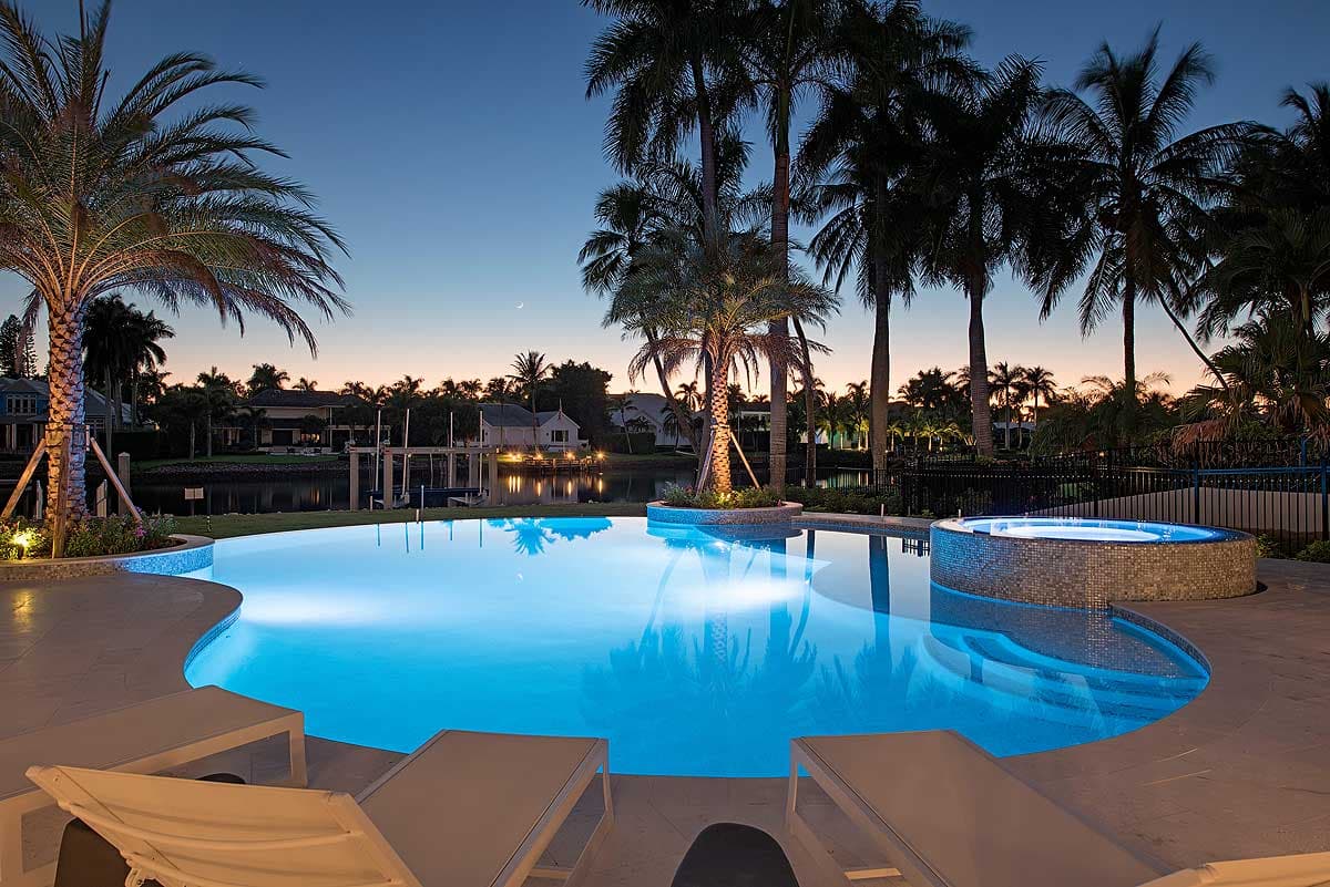 Modern infinity pool with attached illuminated spa, surrounded by palm trees and waterfront homes.