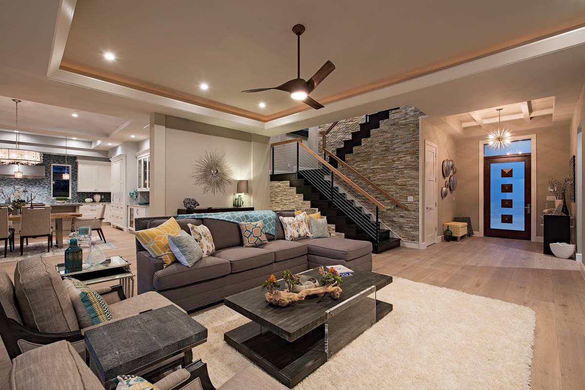 Contemporary living room with sectional sofa, stone accent wall, open staircase, and dining area visible. House plan interior.