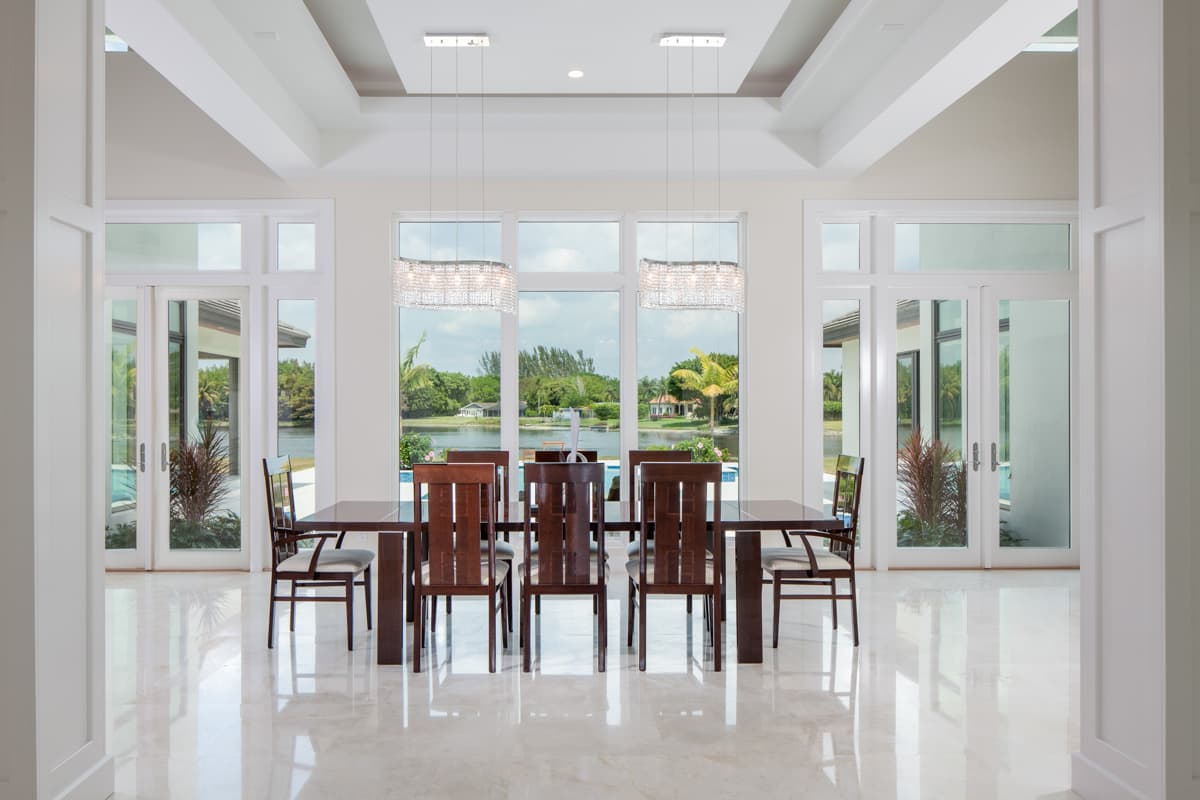 Dining room interior with dark wood table, chairs, and crystal chandeliers overlooking water and landscaping.