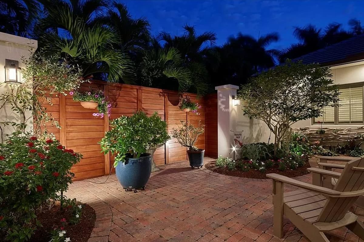 Patio with brick pavers, wooden fence, potted plants, seating, and exterior lighting.