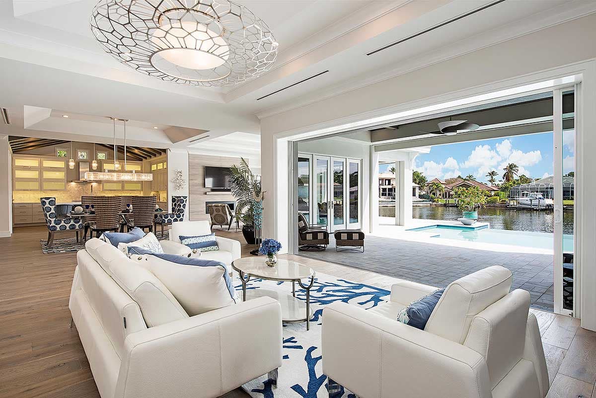 Open-concept living area with white sofas, large windows, dining area, kitchen, and pool view.
