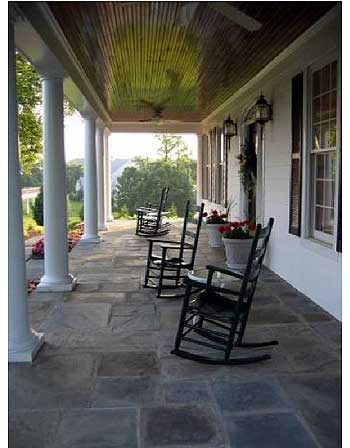 House plan exterior showing a wide covered porch with stone flooring, white columns, and rocking chairs. The ceiling is paneled.