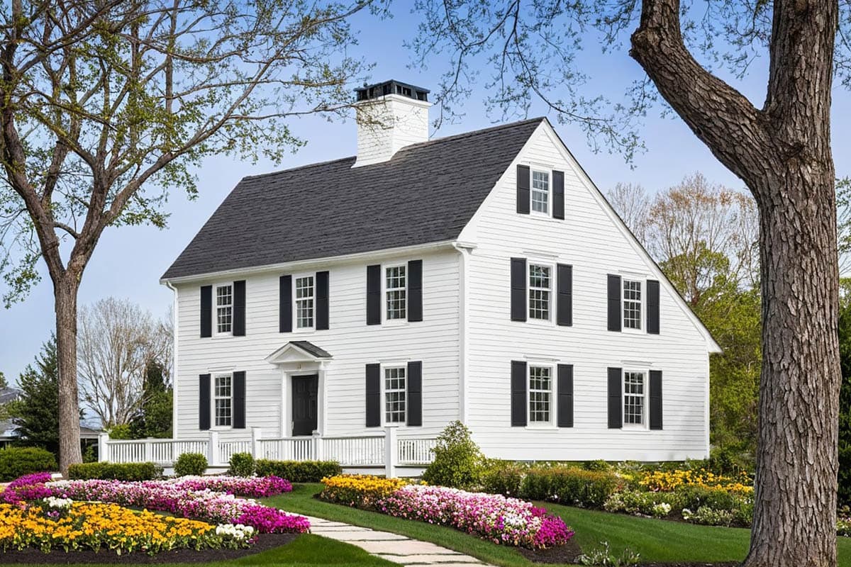 House plan exterior: Two-story white clapboard home with dark shutters, a dormer, and a prominent chimney.