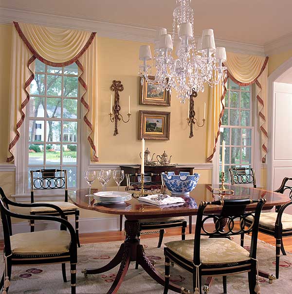 Dining room with oval wood table, upholstered chairs, crystal chandelier, and large windows with draped curtains.